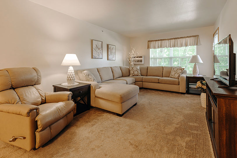 A cozy living room with beige carpet, a large sectional sofa, a recliner, two end tables with lamps, wall art, a window with curtains, and a wooden TV stand with a flat-screen TV.