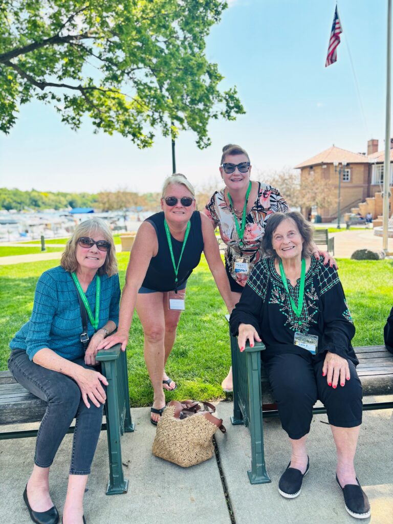 Four women pose and smile outdoors on a sunny day. Three are seated on benches, and one is standing behind them. All wear green lanyards and sunglasses. There’s a marina, grass, and an American flag in the background.