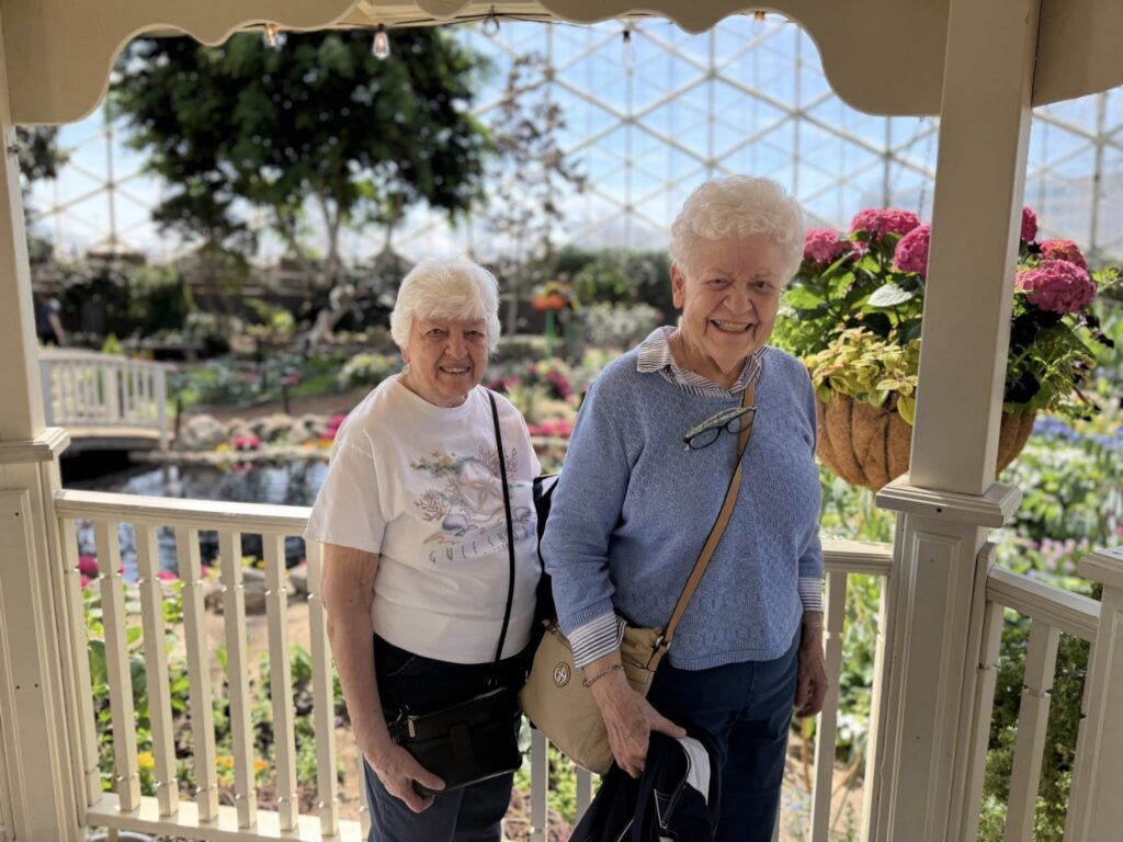 Two elderly women smiling and standing on a porch in a sunlit greenhouse, surrounded by colorful flowers and greenery, with a hanging basket of pink flowers in the foreground.