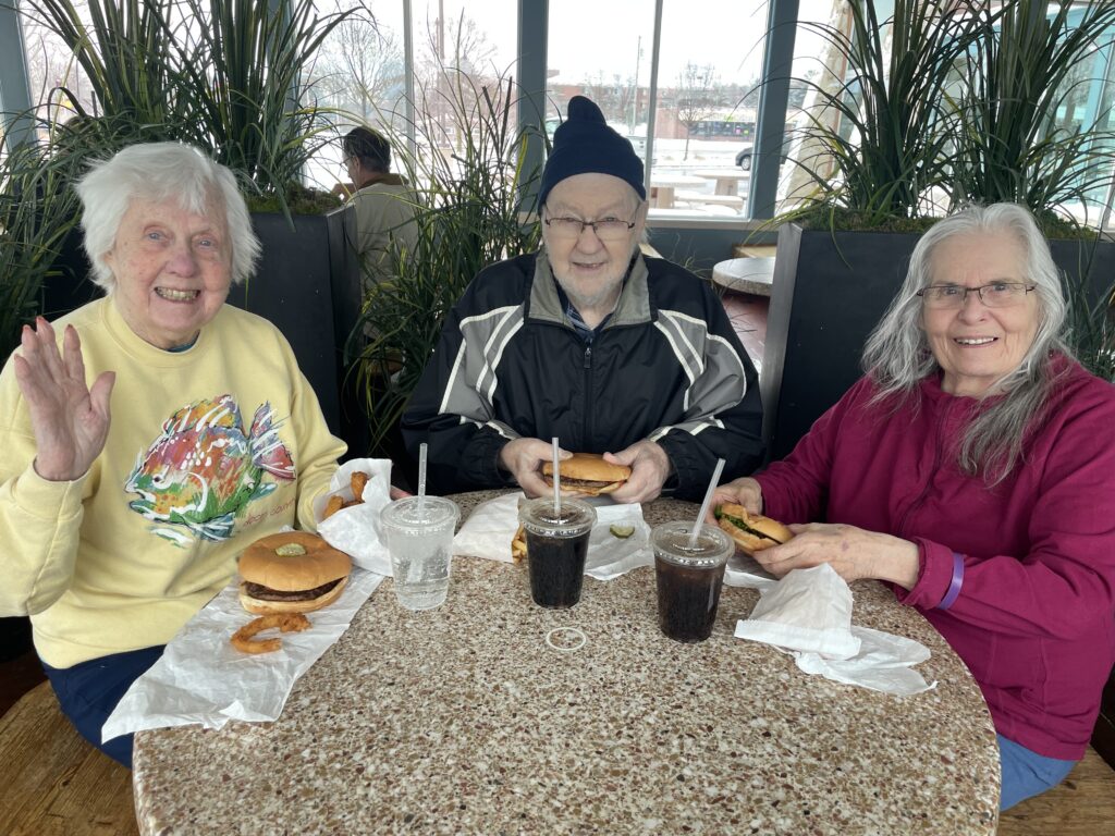 Three elderly people sit at a round table in a restaurant, smiling and enjoying burgers, fries, and drinks. One woman waves at the camera. Green plants and large windows are in the background.