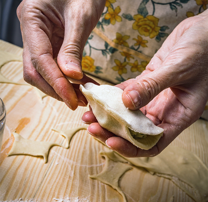 Two hands are folding a piece of dough with a filling inside, making a dumpling. There are pieces of rolled-out dough and flour scattered on a wooden surface in the background.