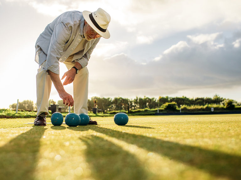 An older man wearing a hat, light shirt, and pants arranges blue lawn bowls on a grassy field under a cloudy sky, with long shadows cast on the ground.
