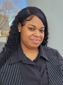 A woman with long, wavy black hair wearing a black shirt and a pinstriped vest stands outside, smiling slightly. Leafless trees and a building are visible in the blurred background.