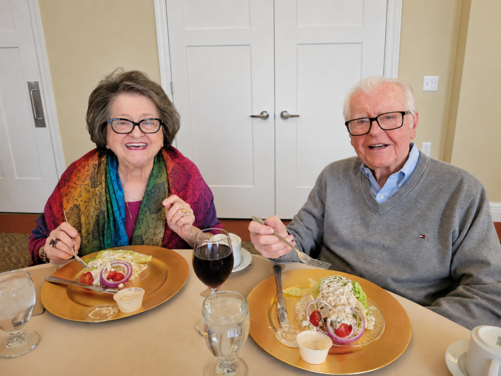 An older woman and man sit at a table, smiling, with plates of salad, rolls, and drinks in front of them. The woman wears glasses and a colorful scarf; the man wears glasses and a gray sweater.