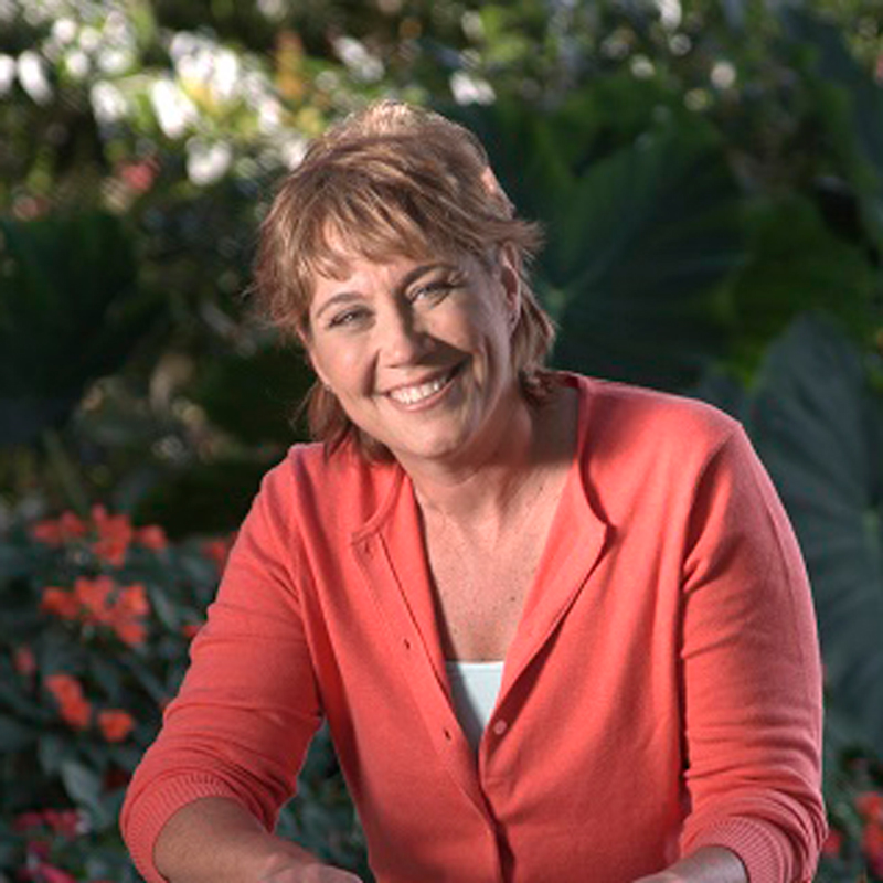 A woman with short light brown hair, wearing a coral cardigan and white top, smiles while sitting outdoors with green foliage and orange flowers in the background.