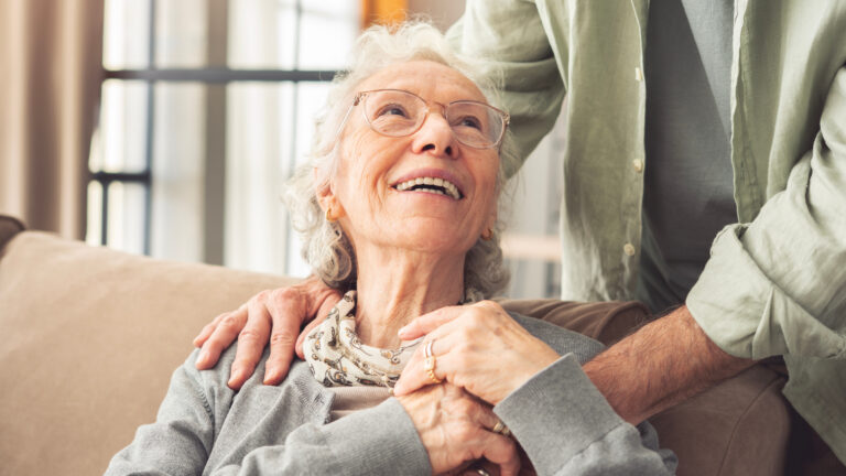 An elderly woman with gray hair and glasses smiles warmly while sitting on a couch, holding hands with someone standing beside her. She looks up at the person with a joyful expression.