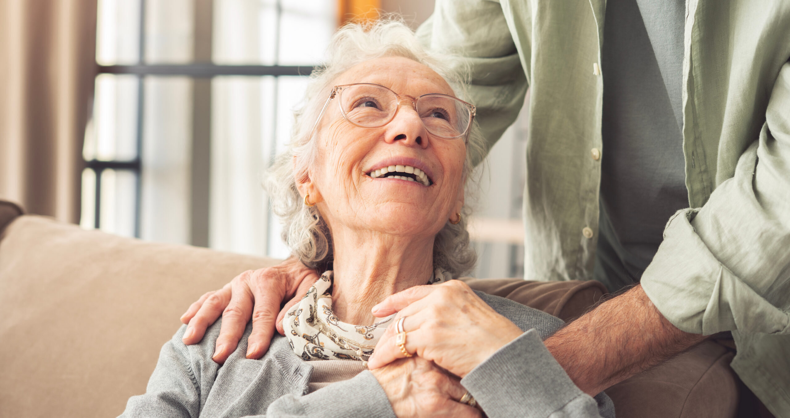 An elderly woman with gray hair and glasses smiles warmly while sitting on a couch, holding hands with someone standing beside her. She looks up at the person with a joyful expression.