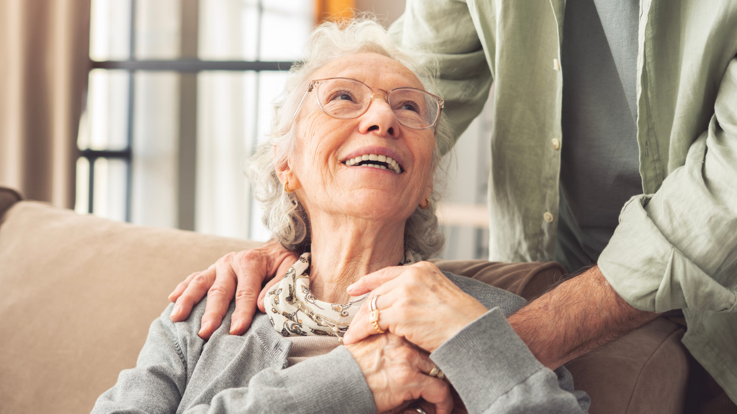 An elderly woman with gray hair and glasses smiles warmly while sitting on a couch, holding hands with someone standing beside her. She looks up at the person with a joyful expression.