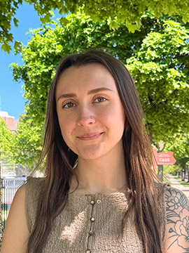 A woman with long brown hair and a sleeveless beige textured top stands outdoors, smiling softly. Green trees and a sunny blue sky are in the background. Part of a tattoo is visible on her upper arm.