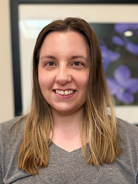 A woman with straight, shoulder-length brown hair and a gray shirt smiles at the camera. There is a blurred framed artwork with purple flowers in the background.
