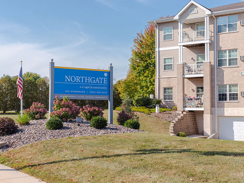 A large blue and yellow sign reading “NORTHGATE: A Capri Community” stands in front of a beige apartment building, with landscaped bushes, an American flag, and a clear blue sky in the background.