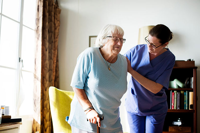 A nurse in blue scrubs assists an elderly woman with white hair and glasses, who is using a cane, as she stands up from a chair in a well-lit room.