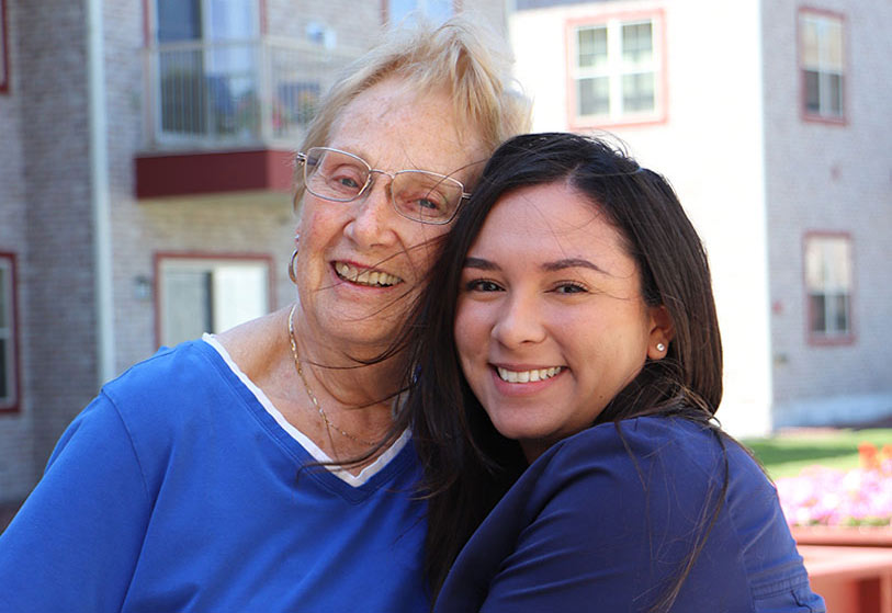 An older woman and a younger woman smile and embrace each other outdoors in front of an apartment building on a sunny day.