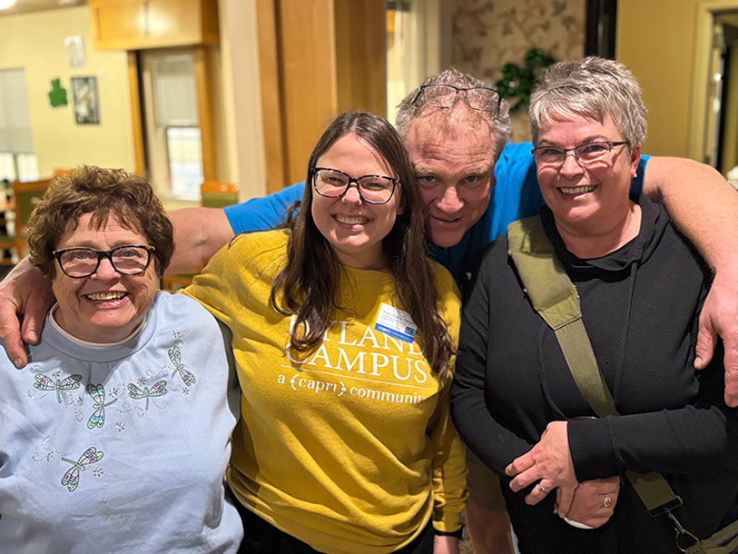 Four smiling adults stand close together indoors, with one man at the back and three women in front. The woman in the center wears glasses and a yellow sweatshirt. They appear happy and are posing for the photo.