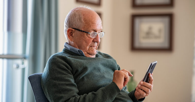 An elderly man wearing glasses and a green sweater sits in a chair, looking intently at a smartphone while using his finger to touch the screen. Framed pictures hang on the wall in the background.