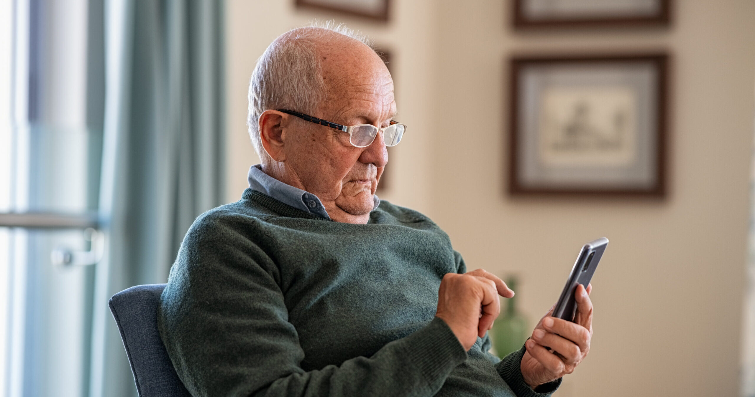 An elderly man wearing glasses and a green sweater sits in a chair, looking intently at a smartphone while using his finger to touch the screen. Framed pictures hang on the wall in the background.