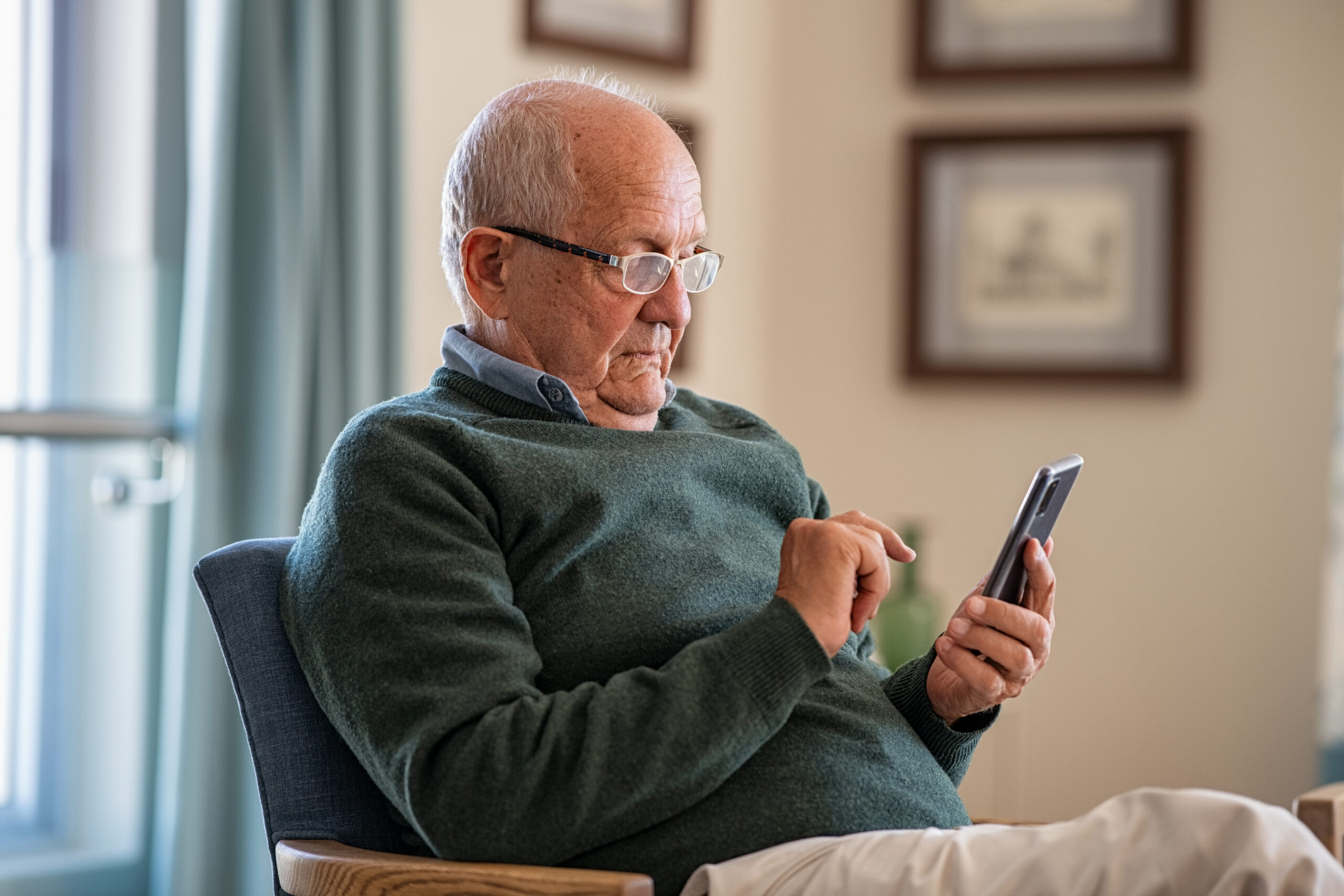 An elderly man wearing glasses and a green sweater sits in a chair, looking intently at a smartphone while using his finger to touch the screen. Framed pictures hang on the wall in the background.