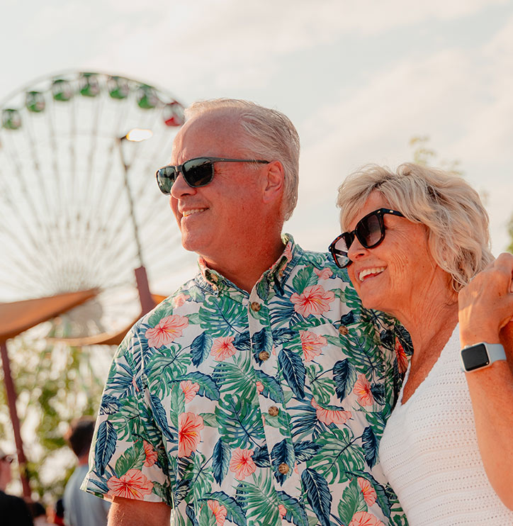 An older couple wearing sunglasses smiles and poses together outdoors, with a Ferris wheel blurred in the background. The man wears a colorful floral shirt, and the woman wears a white top and smartwatch.