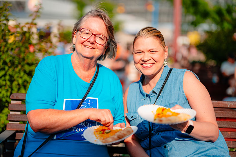Two women sit on a bench outdoors, smiling and holding plates of food. One wears glasses and a blue shirt, the other has her hair pulled back and wears a sleeveless denim top. The background is sunny and blurred.