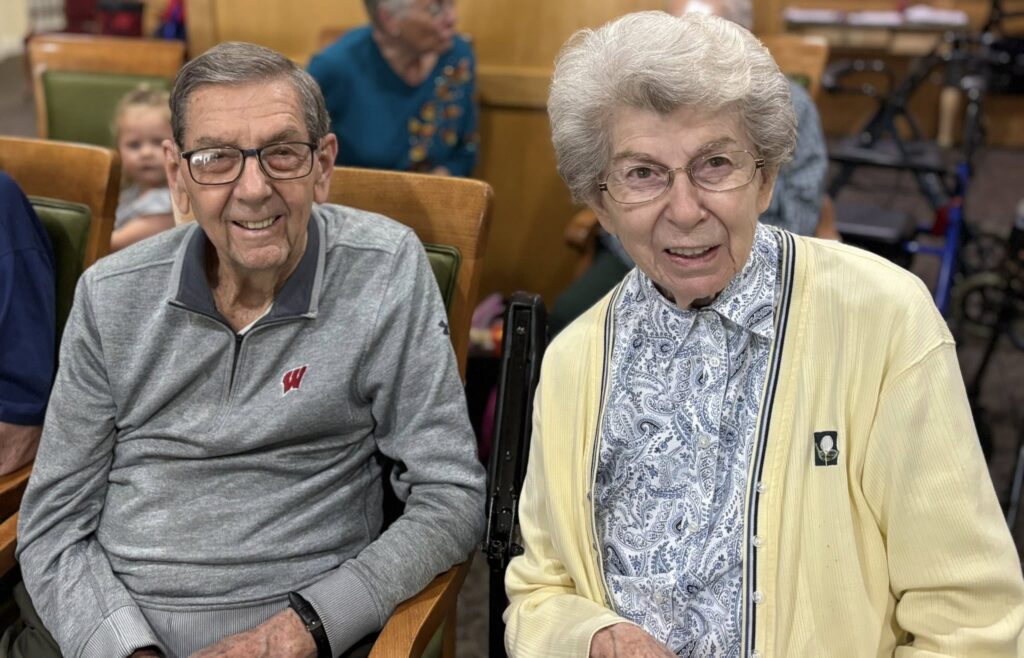 Two elderly people, a man in glasses and a gray shirt with a red W, and a woman in glasses and a yellow cardigan, smile while sitting together indoors. Other people are blurred in the background.