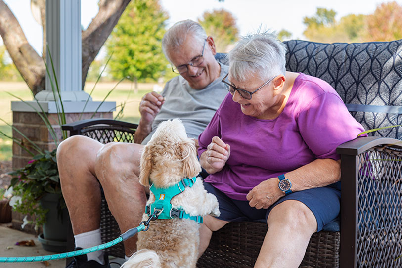 An elderly couple sits on an outdoor bench, smiling and playing with a small, fluffy dog wearing a blue harness. The woman is offering a treat to the dog, while the man watches happily. Trees and greenery are visible in the background.