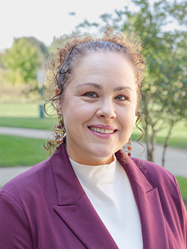 A woman with curly hair wearing a maroon blazer and white turtleneck smiles outdoors, with green trees and grass in the background.