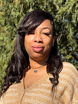 A woman with long dark hair wearing a light brown sweater and a heart-shaped necklace stands outdoors in sunlight, with green trees in the background.