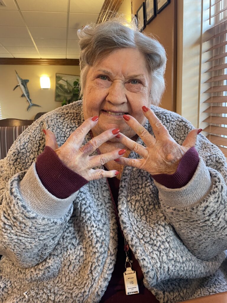 An elderly woman with short gray hair smiles and shows off her freshly painted red fingernails. She wears a cozy gray and maroon jacket and sits indoors, with sunlight streaming through window blinds behind her.