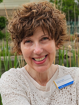 A woman with curly brown hair, wearing a white knit sweater and a name badge, smiles outdoors in front of green plants and a pond.
