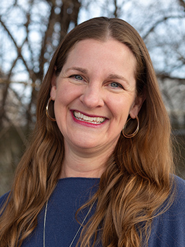 A woman with long brown hair and hoop earrings smiles outdoors, wearing a blue top. Leafless trees and a blurred background are visible behind her.