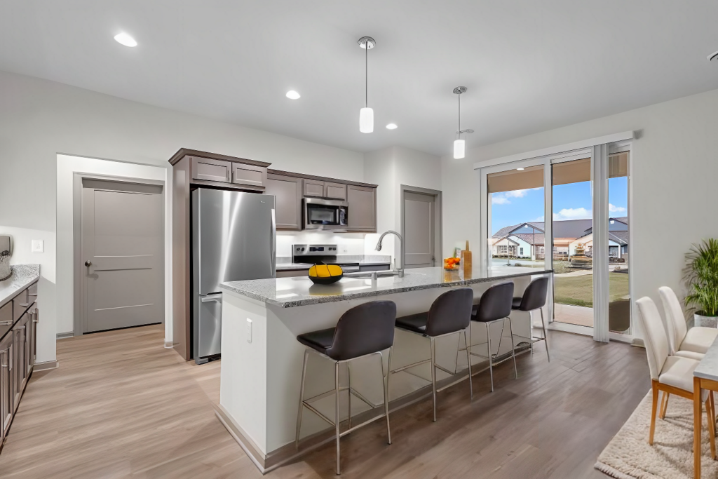 Modern kitchen with gray cabinets, stainless steel appliances, a large island with four black chairs, pendant lights above, and sliding glass doors leading to a backyard with green grass and houses in the distance.