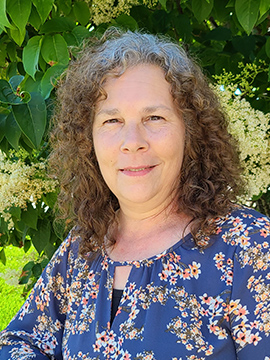 A woman with curly brown hair, wearing a navy blue floral blouse, stands smiling in front of a leafy green background with white flowering plants.