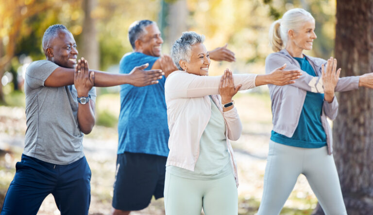 Four older adults exercise outdoors, smiling and stretching their arms across their chests in a park setting, wearing athletic clothing, with trees and greenery in the background.