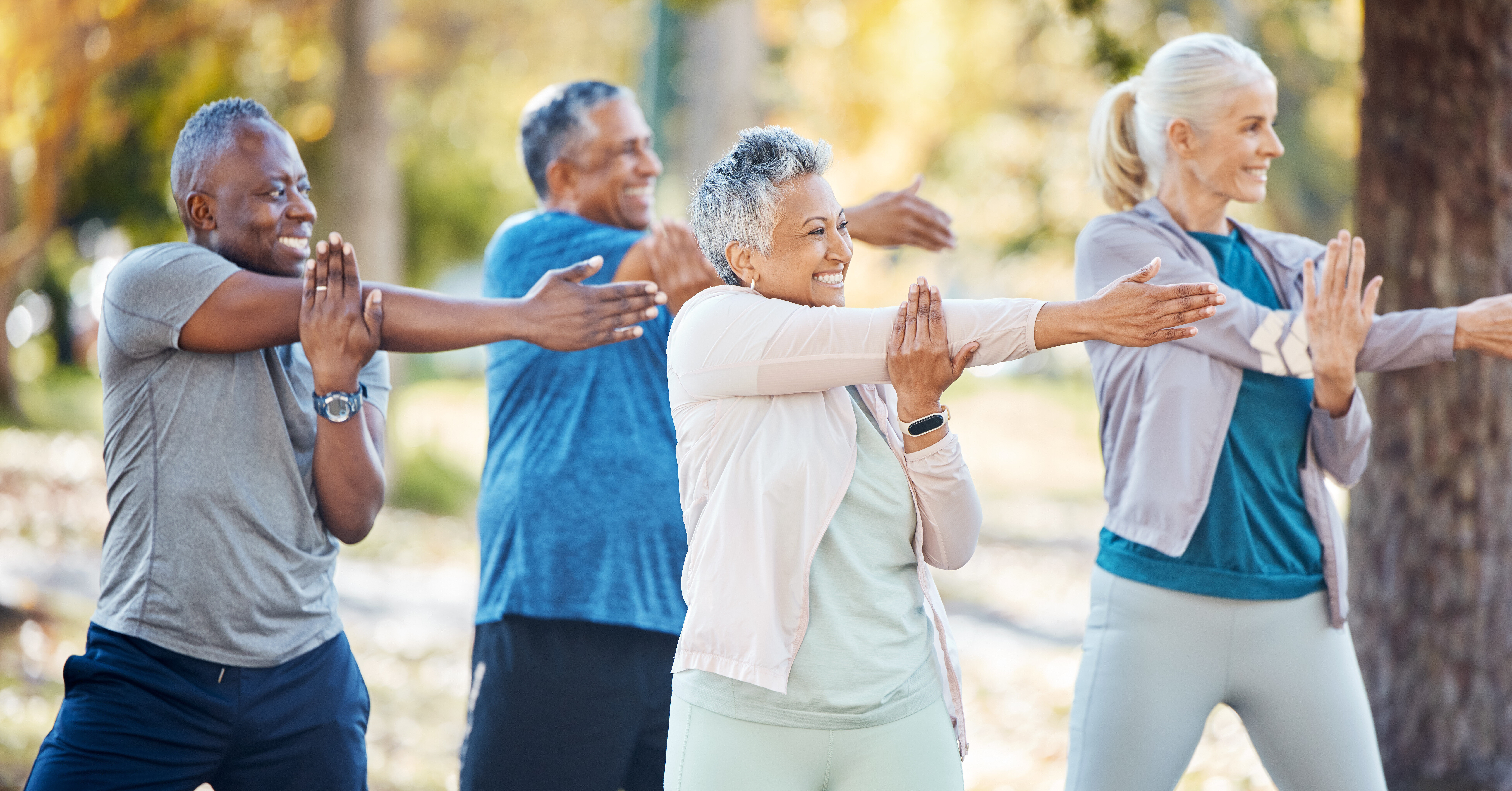 Four older adults exercise outdoors, smiling and stretching their arms across their chests in a park setting, wearing athletic clothing, with trees and greenery in the background.