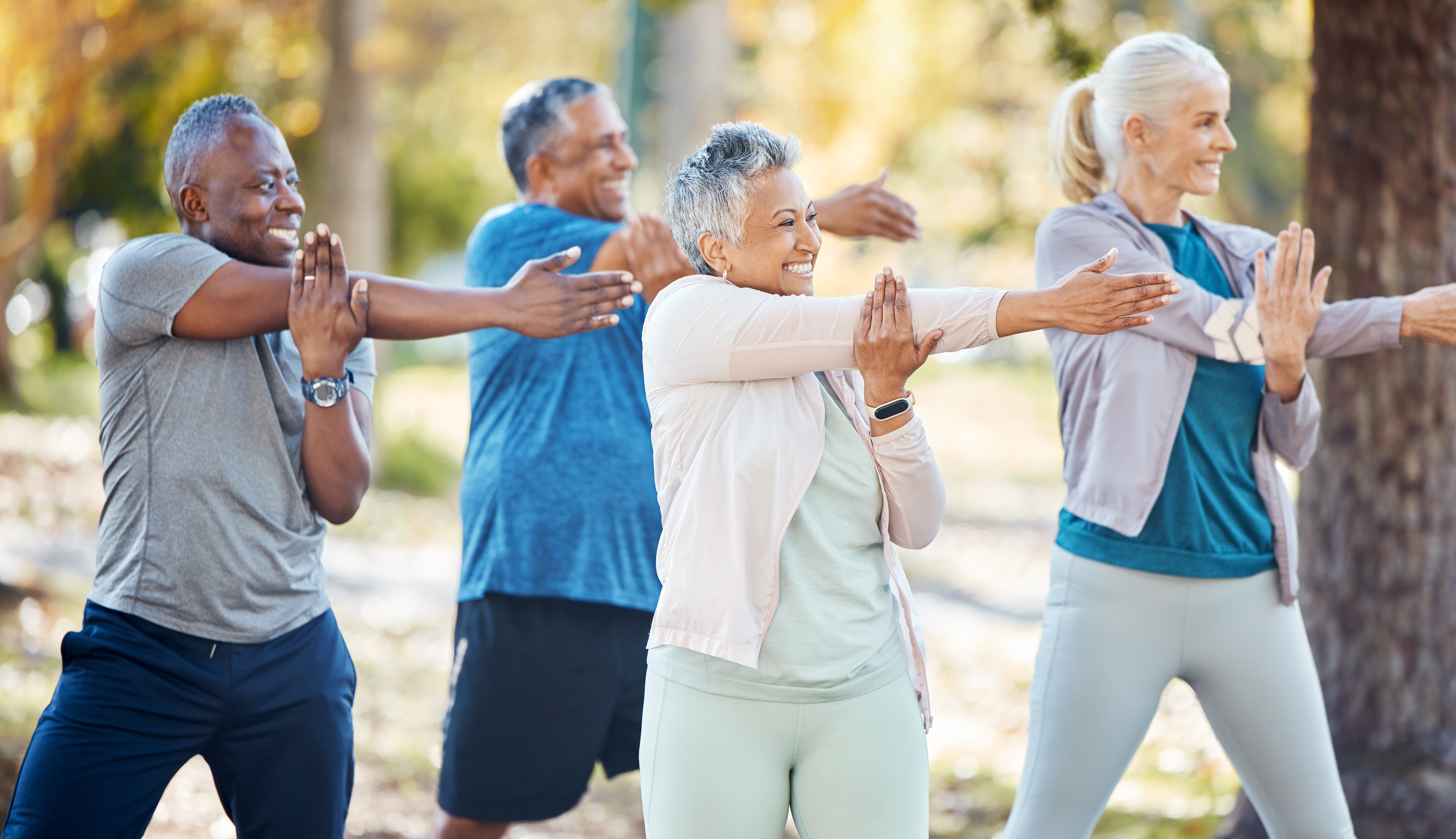 Four older adults exercise outdoors, smiling and stretching their arms across their chests in a park setting, wearing athletic clothing, with trees and greenery in the background.