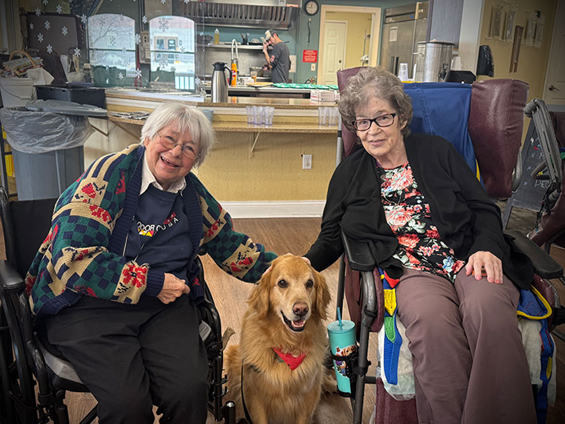 Two elderly women in wheelchairs smile and pet a golden retriever wearing a red bandana. They are indoors, in a cozy kitchen or common area. The atmosphere appears warm and friendly.