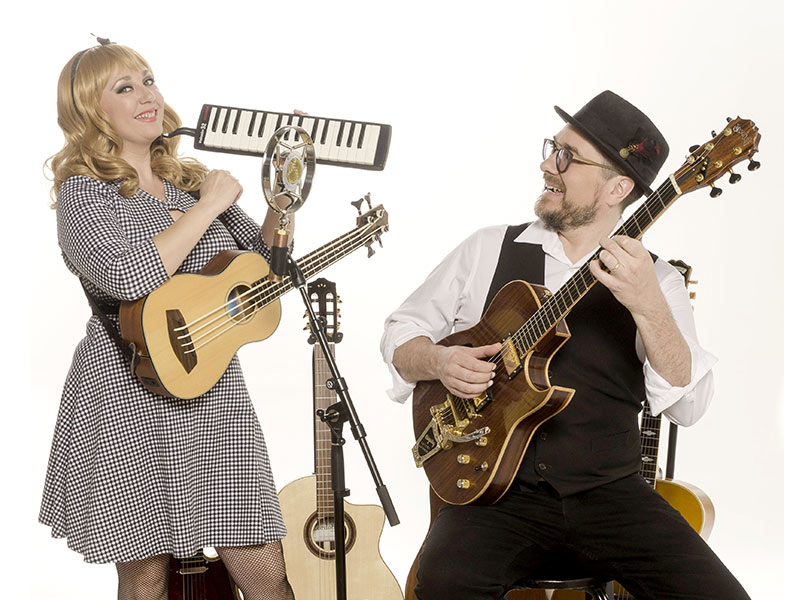 A woman in a checkered dress plays a melodica and holds a ukulele, while a man in a vest and hat plays an electric guitar. Both are smiling, surrounded by several stringed instruments against a white background.
