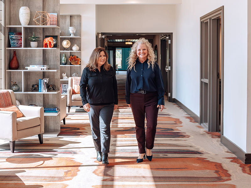 Two women walk together down a hallway with patterned carpet, bookshelves, and comfortable chairs. Both are smiling and dressed in business casual attire in a bright, modern space.