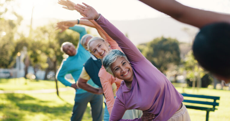 A group of older adults exercising outdoors, standing in a line and stretching with arms raised to the side, smiling and enjoying a sunny day in a park.