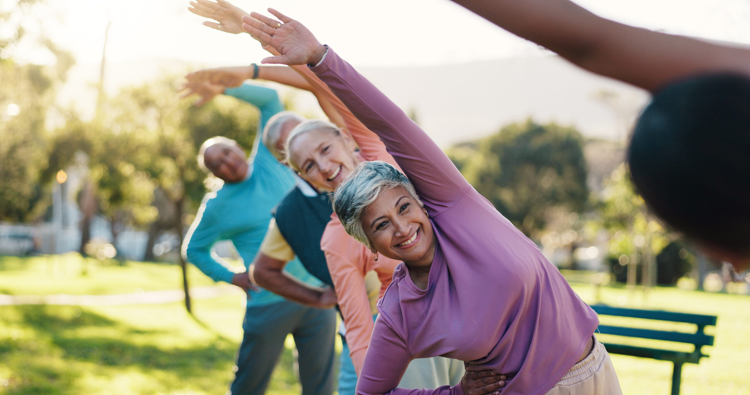 A group of older adults exercising outdoors, standing in a line and stretching with arms raised to the side, smiling and enjoying a sunny day in a park.
