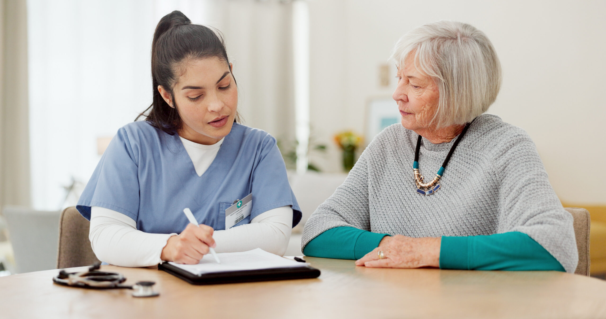 A healthcare worker in scrubs sits at a table, writing on a clipboard, while talking with an older woman who is attentively listening. They appear to be having a consultation in a bright, indoor setting.
