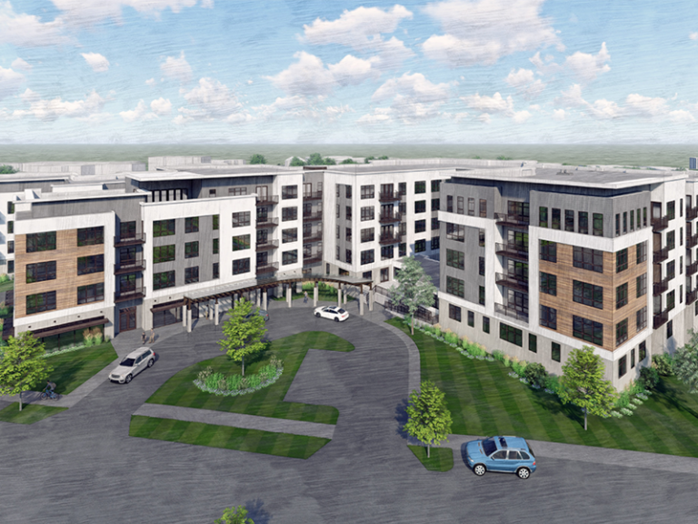 A modern, multi-story apartment complex with white and brown exterior panels, large windows, and balconies. Cars are parked in a circular driveway, surrounded by green lawns, trees, and landscaped areas under a blue sky with clouds.