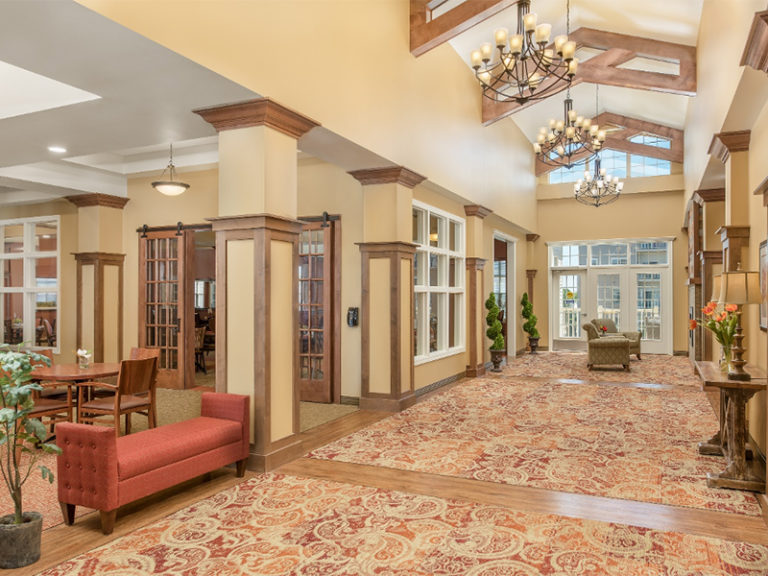 A spacious, elegant lobby with patterned carpet, beige walls, wood trim, large windows, chandeliers, a red upholstered bench, indoor plants, and a seating area in the background. Natural light fills the space.