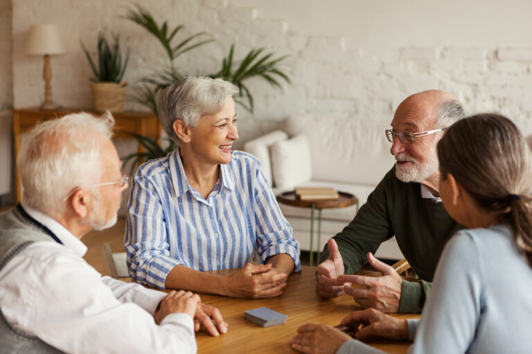 Four older adults sitting around a wooden table, engaged in conversation and smiling. The setting is a cozy, well-lit room with plants and books in the background.