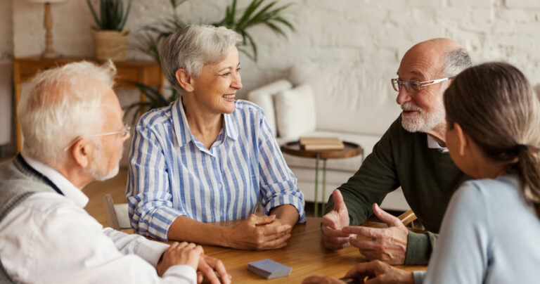 Four older adults sitting around a wooden table, engaged in conversation and smiling. The setting is a cozy, well-lit room with plants and books in the background.