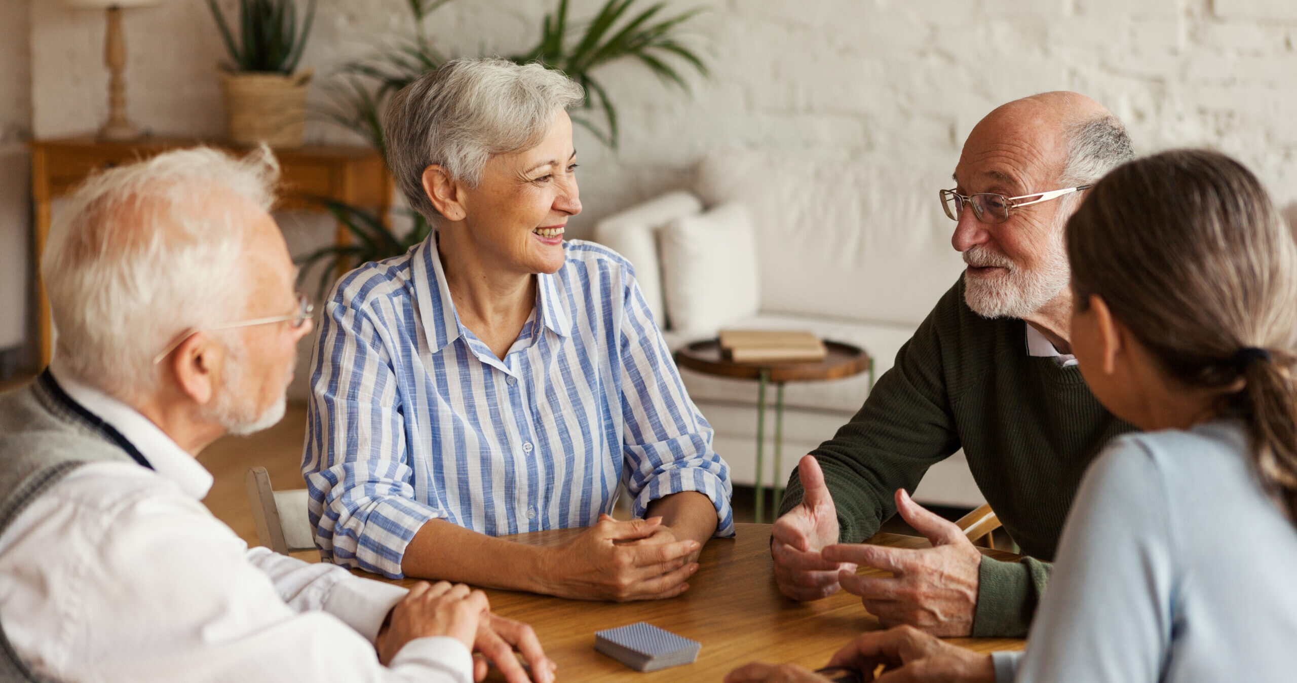 Four older adults sitting around a wooden table, engaged in conversation and smiling. The setting is a cozy, well-lit room with plants and books in the background.