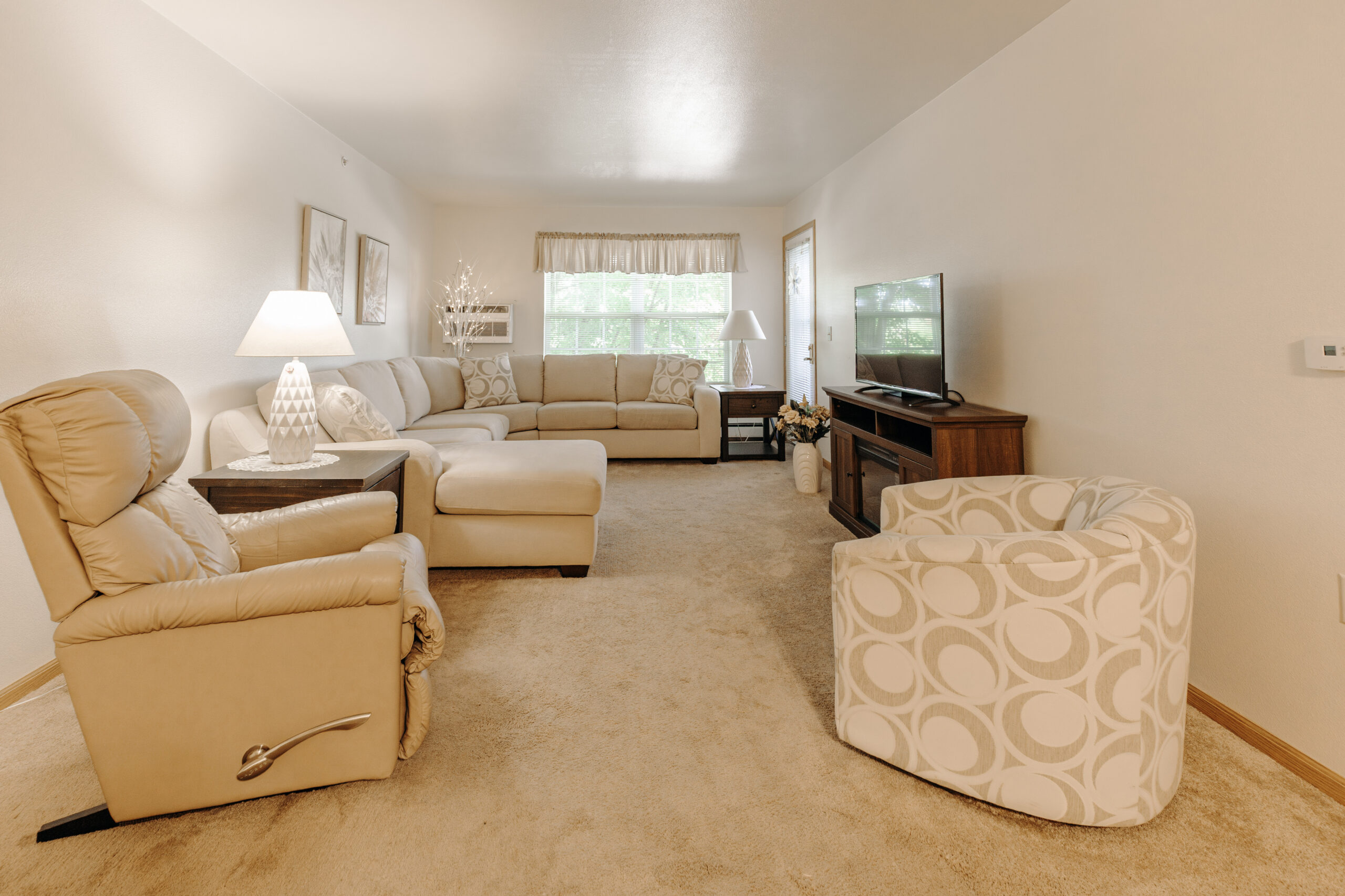 A cozy living room with beige carpet, a sectional sofa, recliner, patterned armchair, a wooden TV stand with a flat-screen TV, side tables with lamps, wall art, and large windows with sheer curtains.