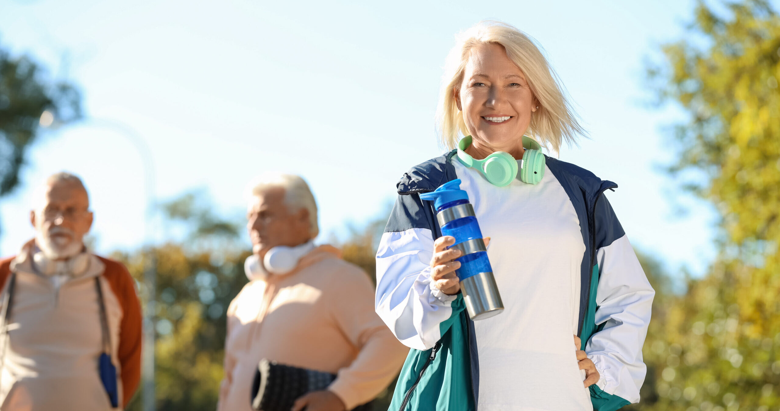 A smiling older woman in activewear holds a water bottle and wears headphones around her neck. Two older men in sports clothes stand blurred in the background outdoors on a sunny day.