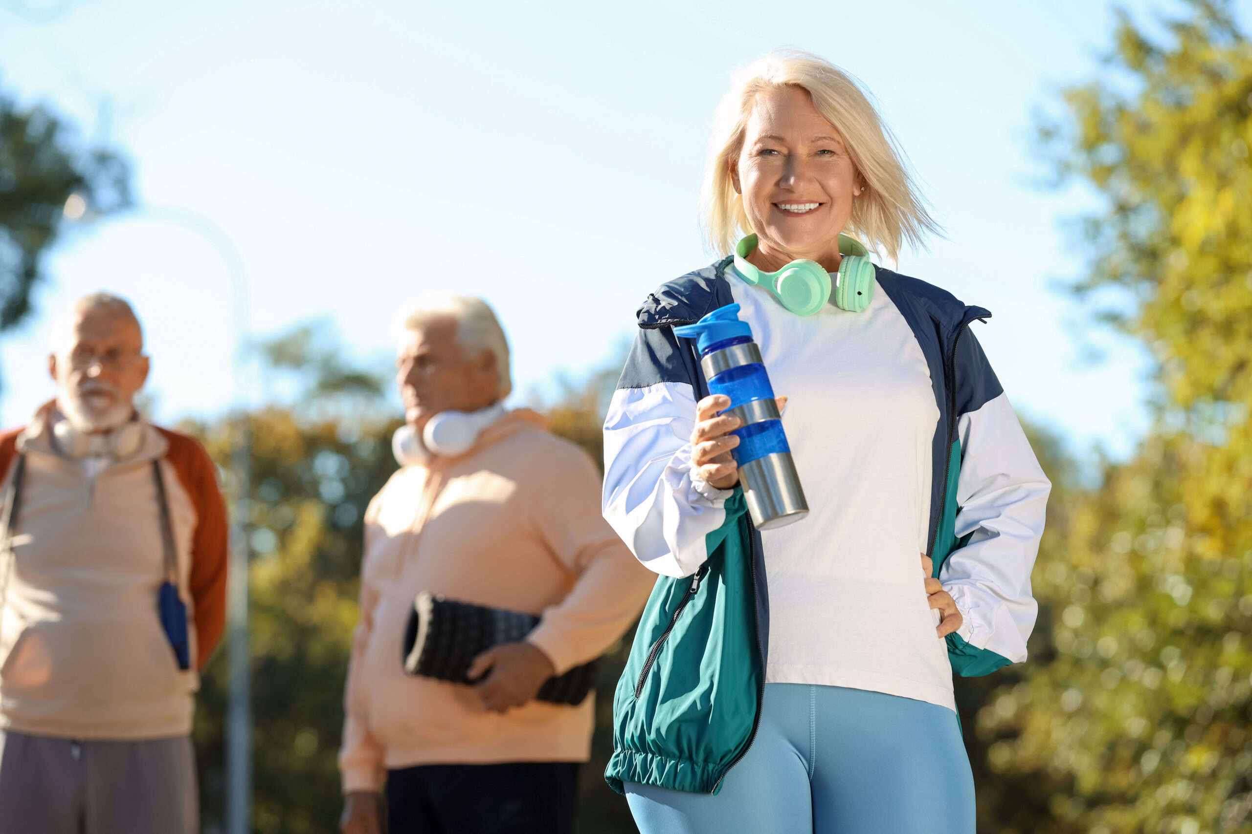 A smiling older woman in activewear holds a water bottle and wears headphones around her neck. Two older men in sports clothes stand blurred in the background outdoors on a sunny day.