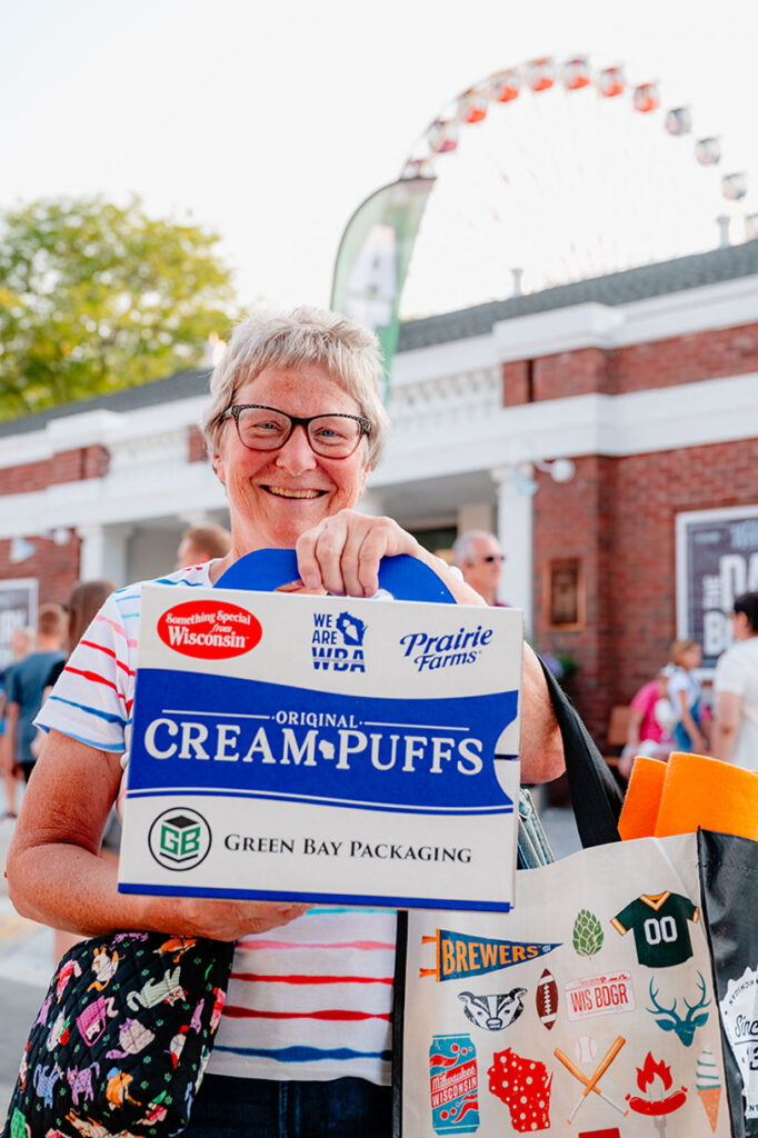 Smiling older woman holding a box of cream puffs at an outdoor event, with a Ferris wheel and brick building in the background. She has short gray hair, glasses, and a tote bag decorated with colorful stickers.